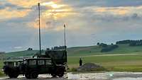 Bundeswehrfahrzeuge mit hohen Masten stehen im Vordergrund vor grünen Hügeln in der Abenddämmerung.