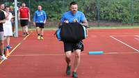 Ein Mann in blauer Sportbekleidung hält vor seinem Körper einen Sandsack mit beiden Händen und läuft.