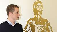 Der Übersetzer Benjamin Weise steht neben einem Wandbild der Star-Wars-Figur C3PO.