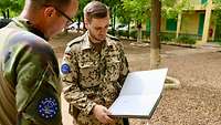 Ein deutscher und ein irischer Soldat stehen in einem gekiesten Innenhof und blicken in ein großes Buch
