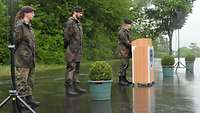 Zwei Soldaten stehen bei Regen neben einem Rednerpult, an dem ein dritter Soldat spricht.