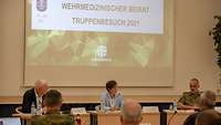 Drei Personen sitzen an Tischen vor einem Publikum