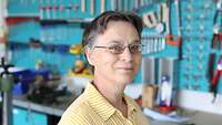 A woman with glasses and a yellow shirt in front of a wall with various tools