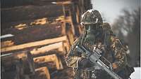 Ein Soldat mit Waffe und ABC-Maske läuft durch den Wald, neben ihm aufgestapelte Holzstämme.