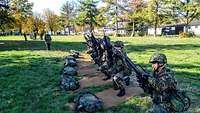 Mehrere Soldaten hocken auf Bastmatten mit ihren Waffen nebeneinander.