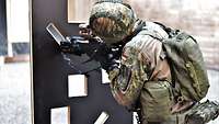 Soldat hockt in unbequemer Haltung vor einem Hindernis und schießt mit dem Gewehr durch eine Öffnung.