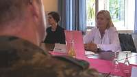 Im Hintergrund sitzen zwei Frauen an einem langen Tisch, im Vordergrund sitzt ein Soldat.