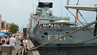 Zahlreiche Menschen gehen einen Kai entlang. Auf der rechten Seite hat ein graues Schiff mit bunten Flaggen festgemacht.