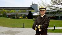 Ein Mann steht in Marine-Uniform in einer parkähnlich angelegten Anlage, im Hintergrund ein großes altes Gebäude