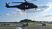 Zwei Soldaten werden gleich eine Palette mit Hilfsgütern an ein Seil hängen, dass zu einem schwebenden Hubschrauber gehört.