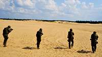 Vier Soldaten laufen nebeneinander mit der Waffe im Anschlag über sandigen Boden 
