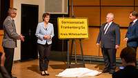 Drei Zivilisten und ein Soldat stehen neben einem gelben Ortsschild, das auf einem Ständer steht.