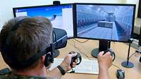 Ein Soldat sitzt mit VR-Brille und Steuerelementen in der Hand vor zwei Bildschirmen.
