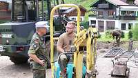 Ein Soldat steht neben einem Minibagger, auf dem ein anderer Soldat sitzt. Beide sprechen miteinander.