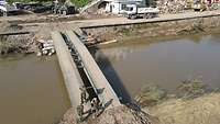Eine militärische Brücke überspannt einen Fluss, im Hintergrund ein betroffener Ort.