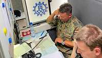 Ein Soldat und ein Mitarbeiter des technischen Hilfswerks telefonieren in einer Kabine.