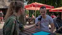 Eine Soldatin und ein Zivilist sitzen sich in einem Biergarten an einem Tisch gegenüber. Sie hält ihm ein Mikrofon hin.