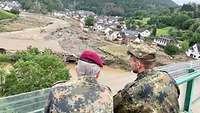 Zwei Soldaten schauen von einer Brücke auf ein vom Hochwasser zerstörtes Dorf