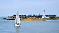 Ein Segelboot segelt vor der Küste, im Hintergrund eine Festung und ein Leuchtturm.