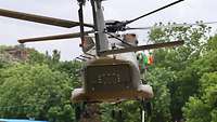 Ein startender Hubschrauber. Im Hintergrund eine malische Flagge und eine rötliche Felswand hinter Bäumen