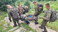 Vier Gebirgsjäger schieben auf einer Militärtrage die verpackte und festgeschnallte Frau auf schmalen Wegen talwärts.