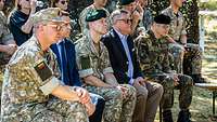 Mehrere Soldaten und zwei Zivilisten im Anzug sitzen auf einer Bänken nebeneinander und lauschen einem Vortragenden.