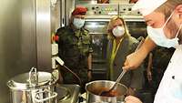 Die Wehrbeauftragte steht neben einem Soldaten und schaut zu, wie zwei Soldaten Suppe vom Topf in eine Schüssel füllen.