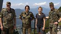 Zwei Soldaten, eine Soldatin und eine Zivilistin stehen auf einer Plattform, im Hintergrund ein Bergpanorama.