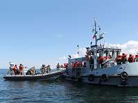 Ein graues Schlauchboot und graues Motorboot nebeneinander in ruhiger See, auf beiden Menschen in Rettungswesten.