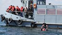 Vor einem grauen Motorboot schwimmt ein Mann in Rettungsweste im Wasser und hebt die Hände.