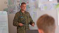 Ein Soldat mit einem Zeigestock steht vor einer Wand mit Kartenmaterial und spricht zu Publikum