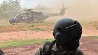 Im Vordergrund ein Soldat mit Fliegerhelm. Im Hintergrund ein Militärhubschrauber der Staub aufwirbelt