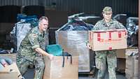 Eine Soldatin trägt einen Karton, ein Soldat schiebt einen großen Karton mit Hilfsgütern