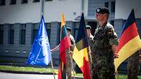 Vier Soldaten stehen mit vier Flaggen in der Hand vor einem Gebäude. Vor ihnen steht ein weiterer Soldat ohne Flagge