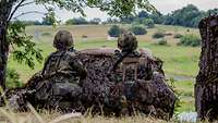 Zwei Soldaten sind mit ihrem Gewehr hinter einem großen Stein in Stellung und schauen in das Gelände vor sich.