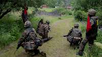 Mehrere Soldaten hocken mit ihren Gewehren auf einem Weg. Zwei Soldaten als Aufsicht stehen mit roten Fähnchen am Rand. 