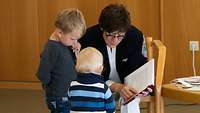 Ministerin Kramp-Karrenbauer sitzt auf einem Stuhl und liest aus einem Buch vor, zwei Kinder stehen vor ihr