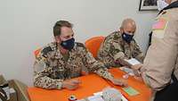 Zwei Soldaten sitzen an einem Tisch. Ein weiterer steht davor und zeigt seine ABC Ausrüstung vor