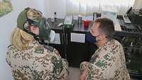 Eine Soldatin sitzt auf einem Stuhl und trägt eine ABC-Maske. Ein weiterer Soldat hockt neben ihr und überprüft die Dichtigkeit.