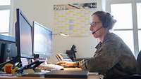 Eine Soldatin sitzt in Uniform mit Headset vor zwei Monitoren an einem Bildschirmarbeitsplatz