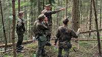 Vier Soldaten stehen im Wald an einem Hang, ein Soldat redet und gestikuliert. 