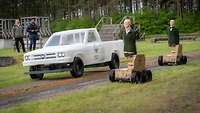 Zwei Puppen auf Rädern und ein weißer Pick-up auf einem Testgelände, im Hintergrund zwei Männer mit Laptops