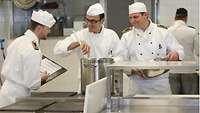Several cooks stand in a canteen kitchen talking to each other.
