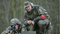 Two soldiers pictured in a wooded area. One of them lies behind a machine gun while the other kneels next to him.
