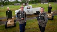 Zwei Männer stehen mit Laptops auf einem Testgelände vor zwei Puppen auf Rädern und einem weißen Pick-up 