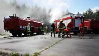 Drei Löschfahrzeuge stehen vor einem rauchenden Hangar. Mehrere Feuerwehrleute laufen dazwischen umher.