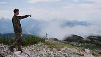 Auf einer Anhöhe steht ein Soldat und zeigt auf die umliegenden Berge, auf denen teilweise Wolken liegen.