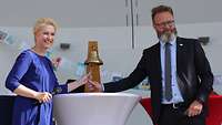 Eine Frau und ein Mann halten ein Bändchen, welche an einer Glocke befestigt ist und lächeln.