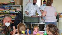 Zwei Köche der Bundeswehr teilen Kindern Mittagessen aus; diese sitzen am Tisch davor