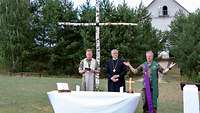Drei Geistliche stehen auf einem Feld vor einem Birkenkreuz bei einem Gottesdienst, vor ihnen ein provisorischer Altar.